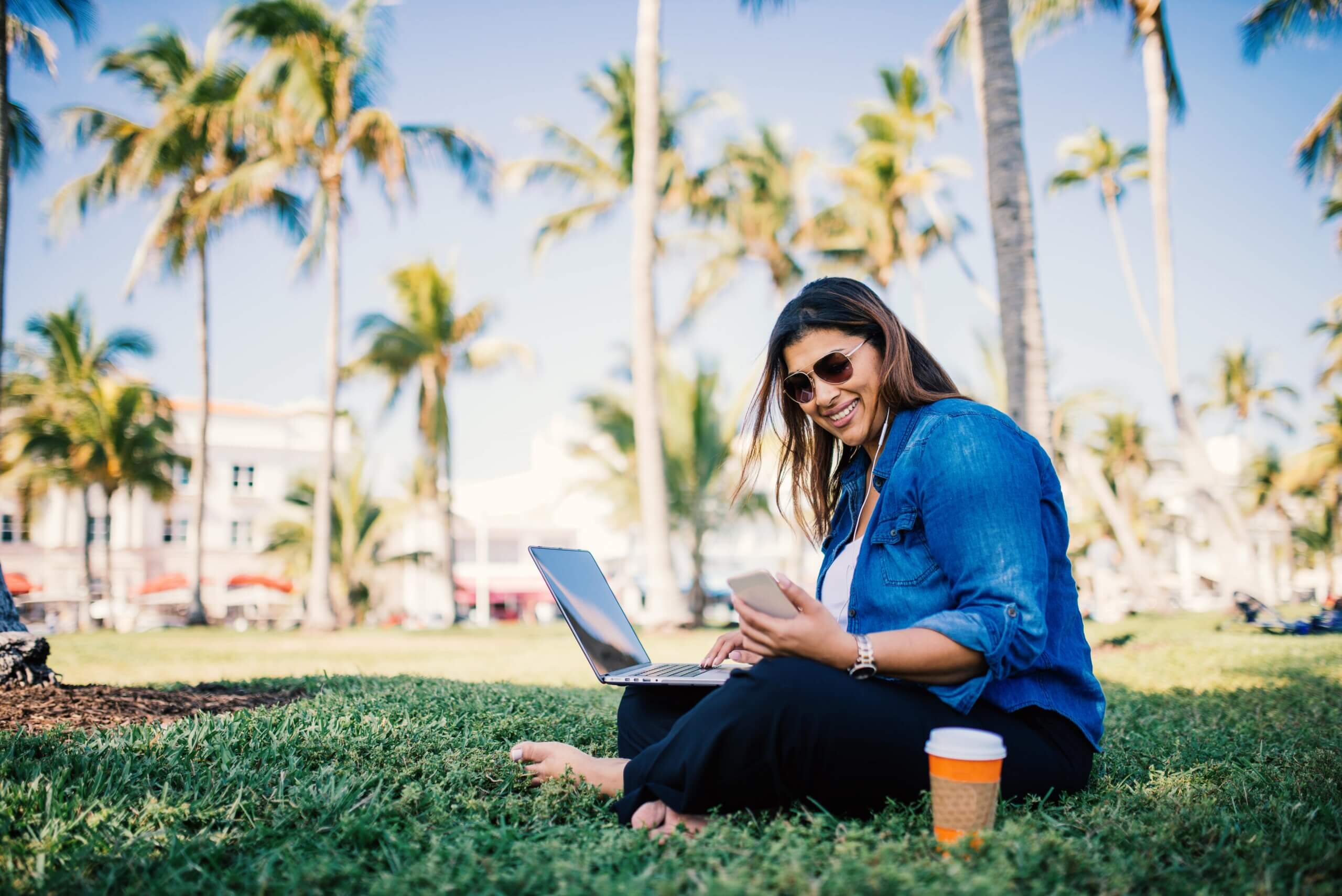 casually dressed woman sitting in the grass in a public park and working on her laptop and phone